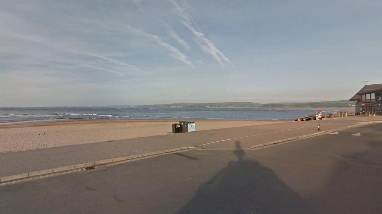 Google Street View of Exmouth beach looking out towards the sea and the sandy beach. A set of three public bins are in the centre of the shot along with the shadow of the Google Street View car.