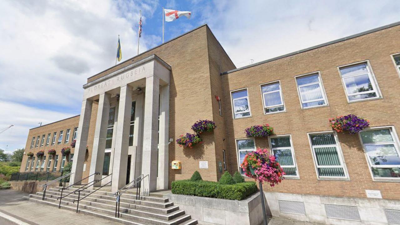 A Google photograph of Rugby Town Hall in Evreux Way. The modern building has steps leading up to the entrance which has pillars and flags flying above it. There are hanging baskets on the front.