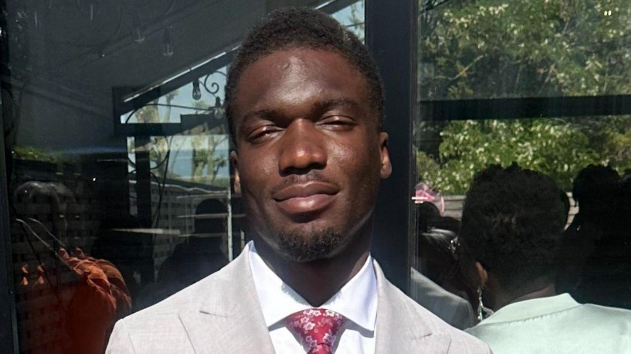 Alex Mamwa wearing a light grey suit and red flowery tie. He is smiling at the camera with the sun shining on his face. 