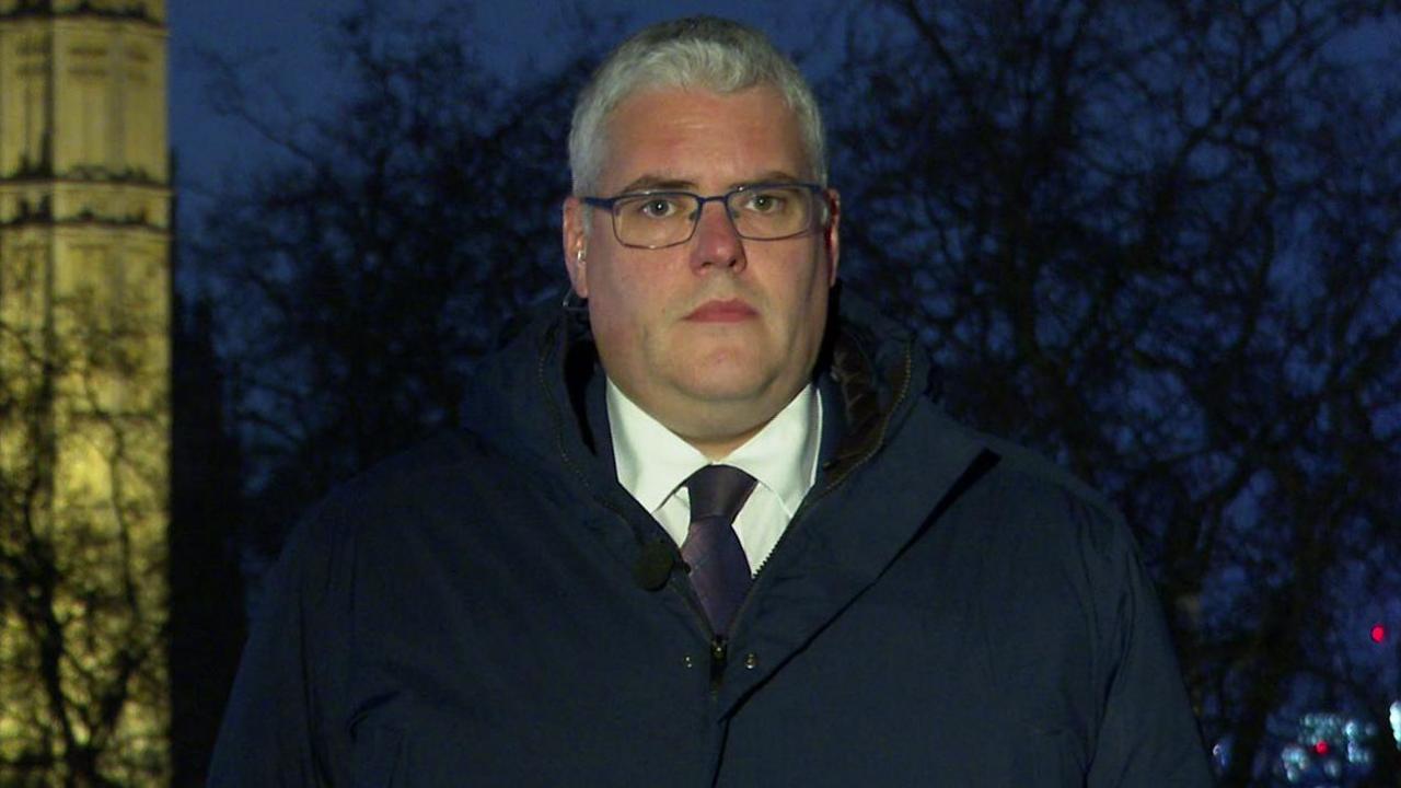 A still of DUP leader Gavin Robinson from the shoulder's up looking directly at the camera. he is wearing a navy coat, a white collared shirt and a dark coloured tie. He is also wearing a pair of metal, rectangle framed glasses. In the background are some trees and a zoomed in portion of Big Ben.