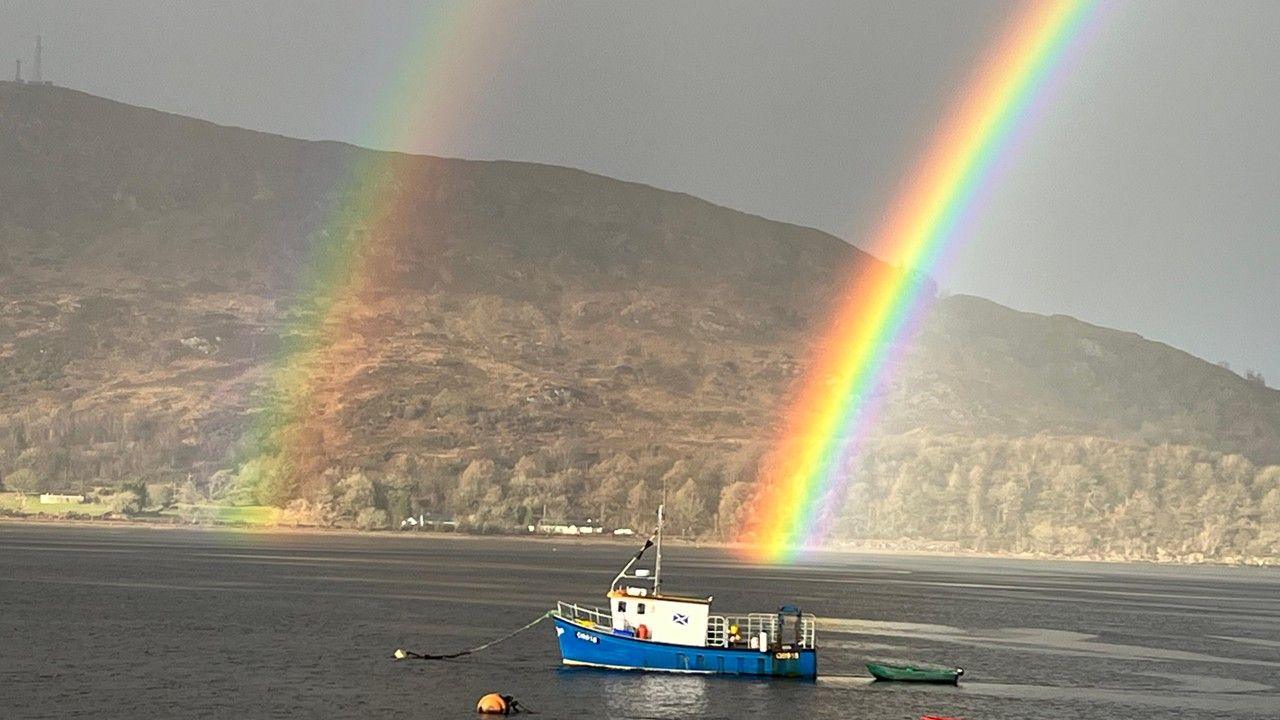 Two rainbows seem to end by the water with a fishing boat in the foreground and mountains behind