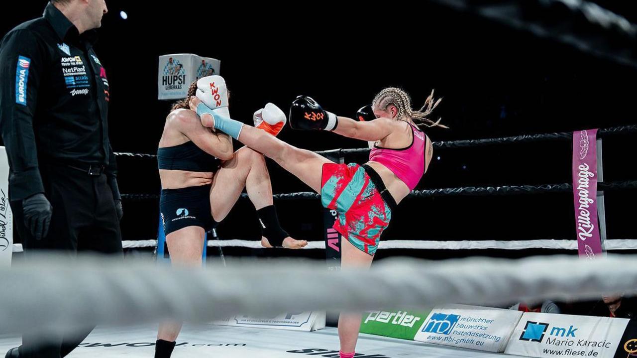 Turner, in pink, landing a high kick on her black-clad opponent in the centre of a boxing ring and a black-clad man watching on the left. A blurred white rope can be seen in the foreground