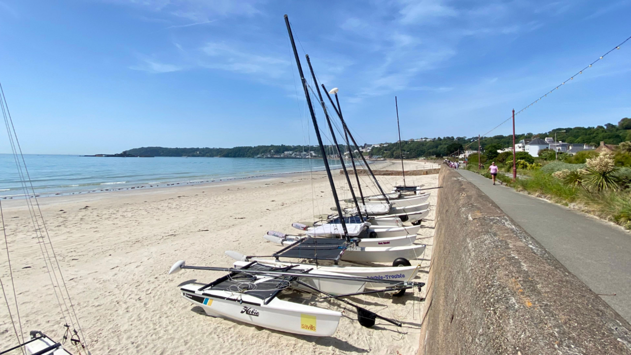 A row of small sailing boats sits on a sandy beach beside a coastal path, with calm blue sea, a distant headland and scattered houses under a bright, lightly clouded sky.