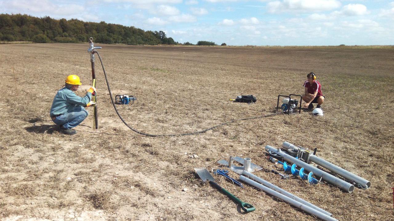 Two people in a large open field which is mostly dirt. They have equipment, including a narrow pipe going into the ground, which is creating a small borehole for sediment samples. To the side, other tools and pipes are laid out. One person is next to a small generator, powering the equipment.