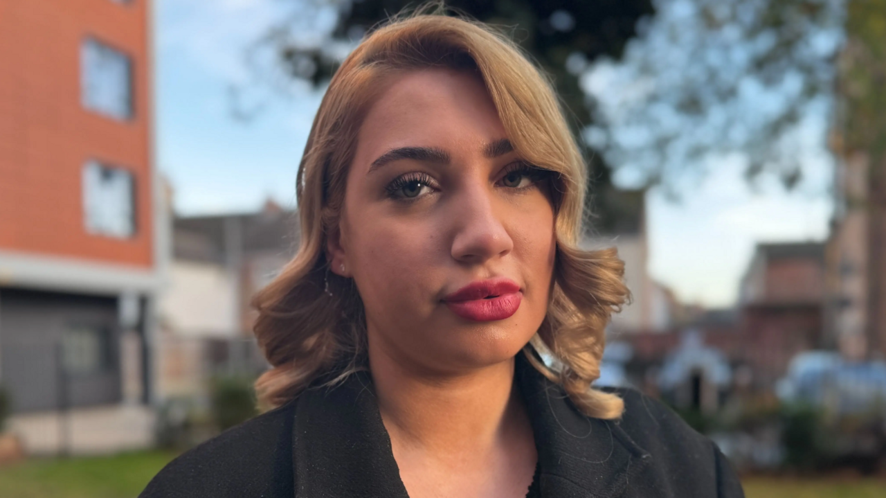 Amira Ostalski is in the middle of the image looking towards the camera. She has shoulder length light brown hair and is wearing a black coat. 