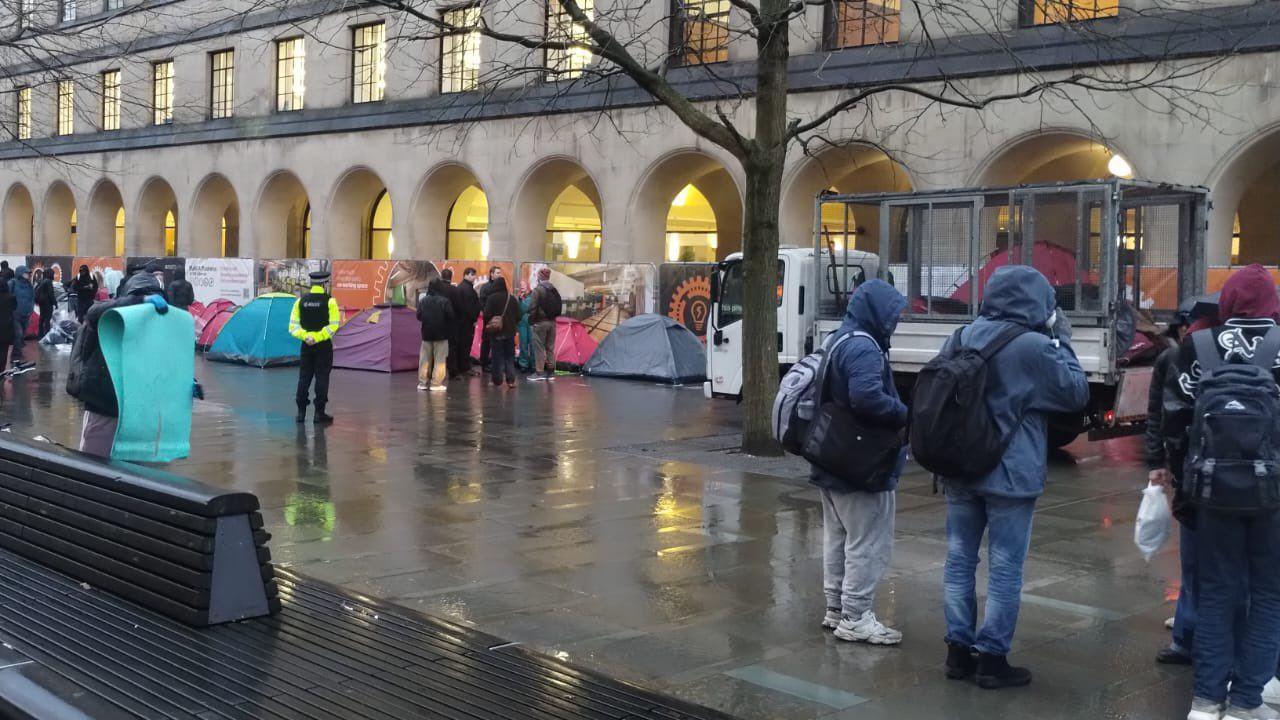 A small van is parked in front of the arches at St Peter's Square as council workers throw red tents into a trailer on the back. Three men wearing hoodies and rucksacks are seen watching on. A group of other people surrounded one of the tents 