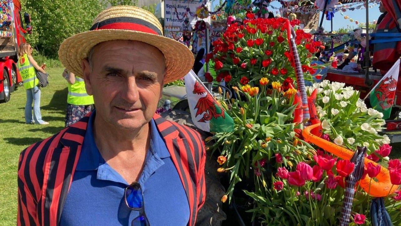 Dai wears a black and red blazer and straw hat with a ribbon. He stands beside his float, which has many tulips displayed in vases.