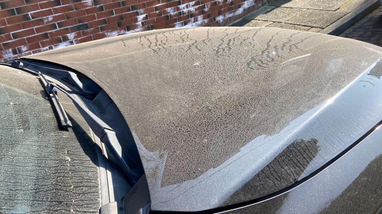 Dust has covered the bonnet of a black car. It is all over the windscreen and there are tracks through the dust on the bonnet.