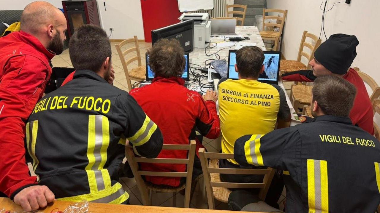 A row of six search and rescue team members in red, black and yellow hi-vis uniforms carefully look over images of the area.