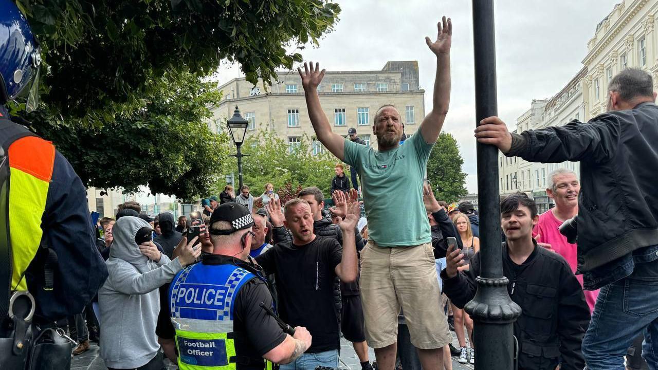 Protesters in Plymouth hold their hands up as police officers watch on. One protester wearing a light green t-shirt and beige shorts is stood on a bench