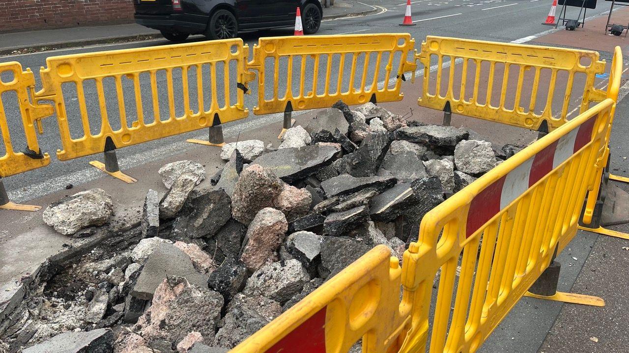 Yellow plastic fencing around a large hole in the bus lane section of Heavitree Road with a pile of rubble filling the hole and overflowing.
