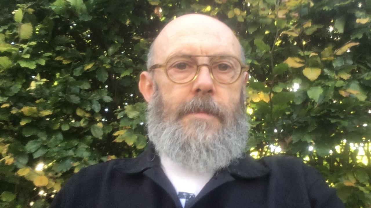 A man wearing glasses with a grey bread. He is outside with and is standing in front of a large hedge 