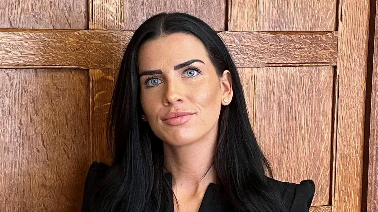 Olivia Gates has long black hair and is wearing a black dress. She is standing in front of a wooden office door.