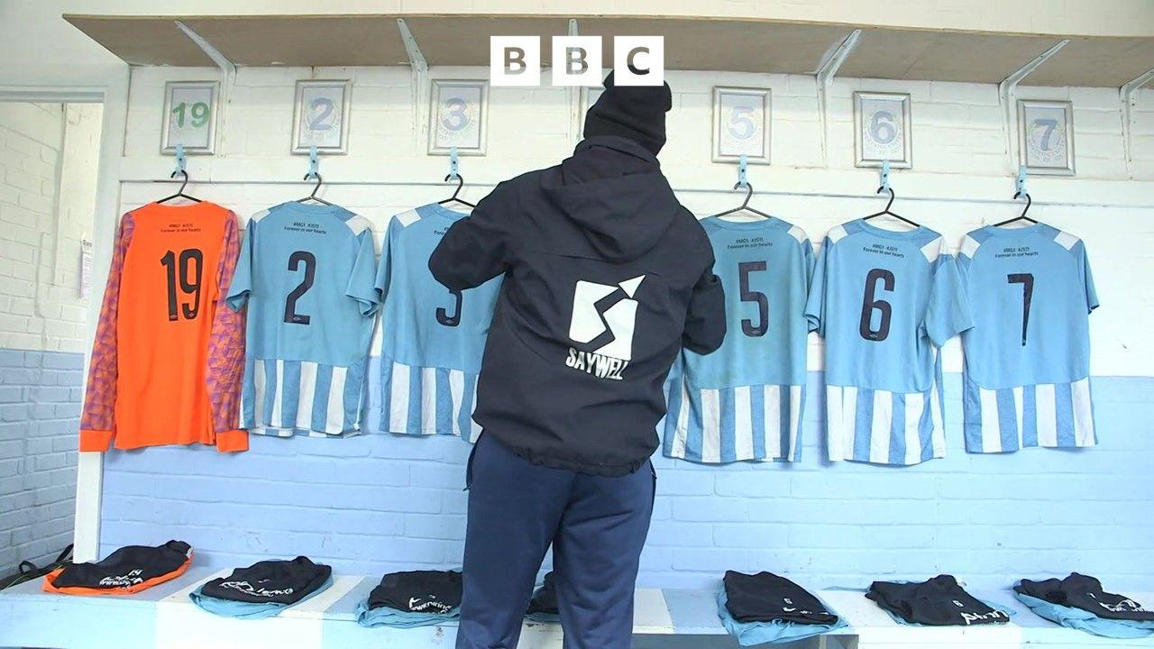 A man hangs pale blue football shirts on hooks in a players dressing room. His back is turned to us. He wears a black hoodie and blue jeans.