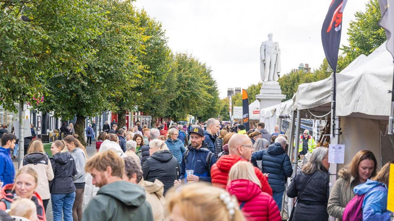 Dozens of people line Cockermouth's Main Street as they browse food stalls. A number of mature trees are growing on both sides of the road and there is a statue in the background to the right.
