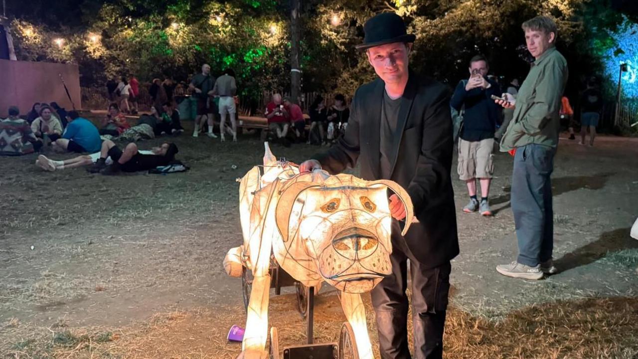 A man in a black suit and hat is standing next to a dog made of paper and lit up from within. Festival-goers can be seen lounging on the floor in the background, with a few stopping to look at the man and his dog lantern.