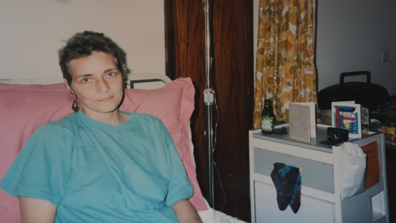 Carolyn Challis laid on a hospital bed during cancer treatment. She is wearing a blue t-shirt and appears to be hooked up to a medical drip. She is looking at the camera.