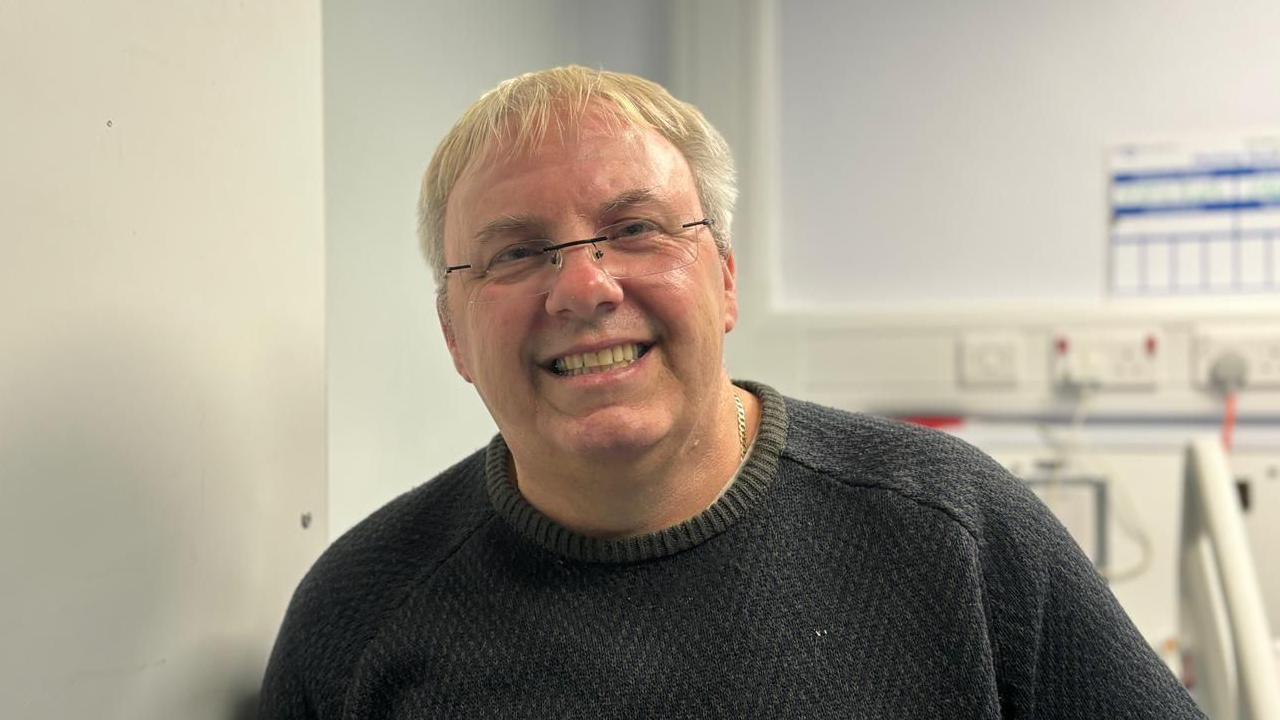 Dave Hunt, a middle-aged blonde man wearing a grey jumper and frameless glasses, leaning on the wall of a hospital ward.