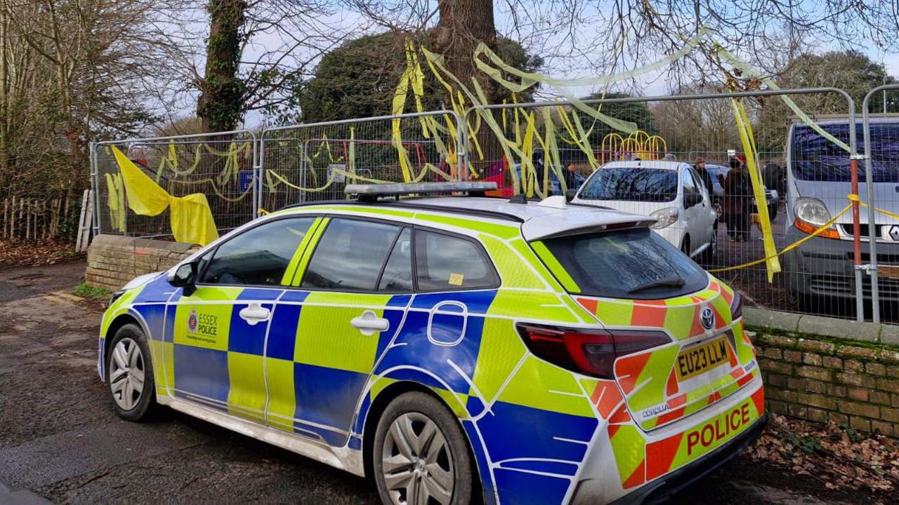 A police car parked in front of an oak tree, which has yellow ribbons attached to its branches. The tree is being held behind heras fencing. Campaigners are stood in the background.