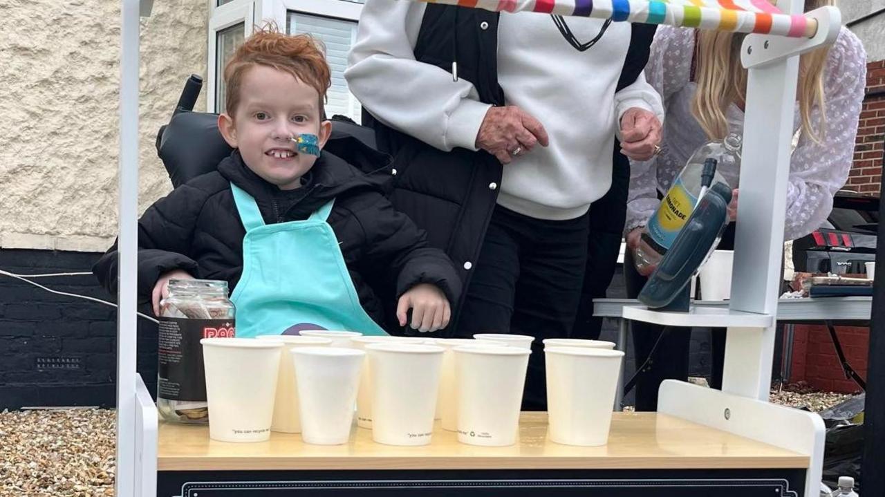 Thomas smiles behind his lemonade stand. Cups of lemonade are on the counter.