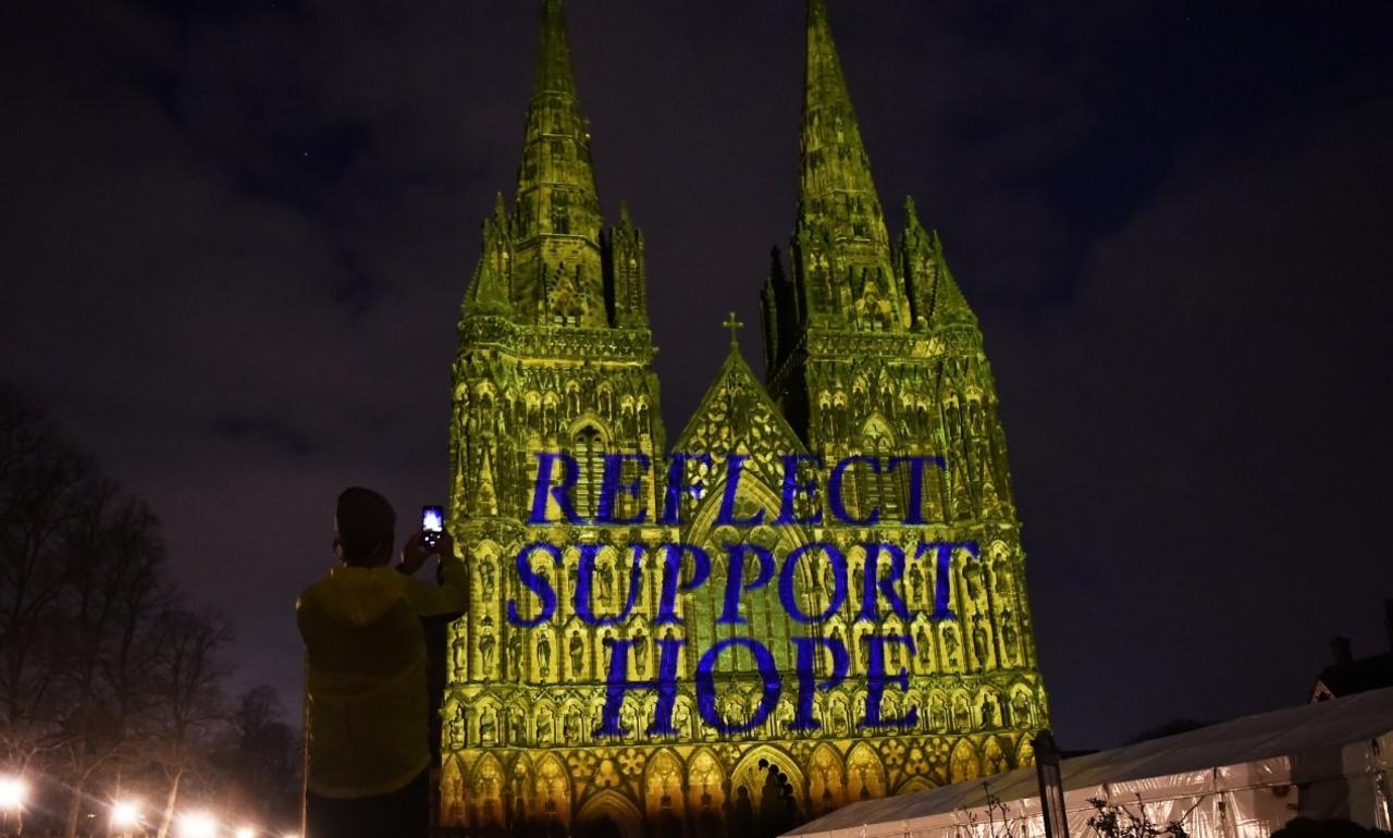 A man takes a photograph of Lichfield Cathedral as it is illuminated in yellow with the words, “Reflect, Support, Hope”.