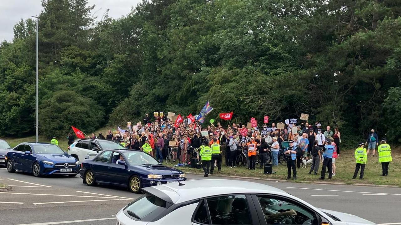A large crowd of people standing by a junction. There is police around the group and protesters are holding flags.