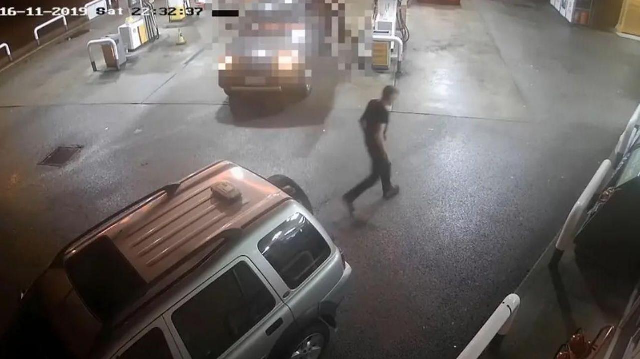 A CCTV camera view shows a person walking across a wet petrol station forecourt at night, with several vehicles parked near the fuel pumps and a timestamp visible in the upper-left corner.