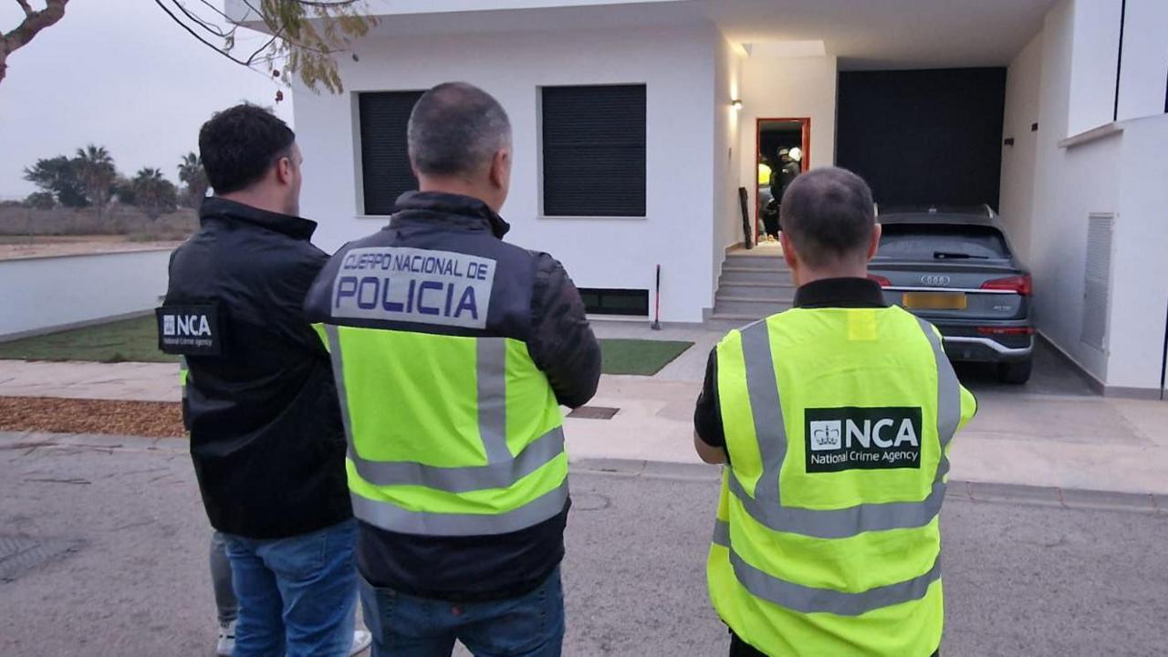 One Spanish Policeman and two NCA officers wearing hi vis at Colin Wright's home in Spain. a white building with one car outside.