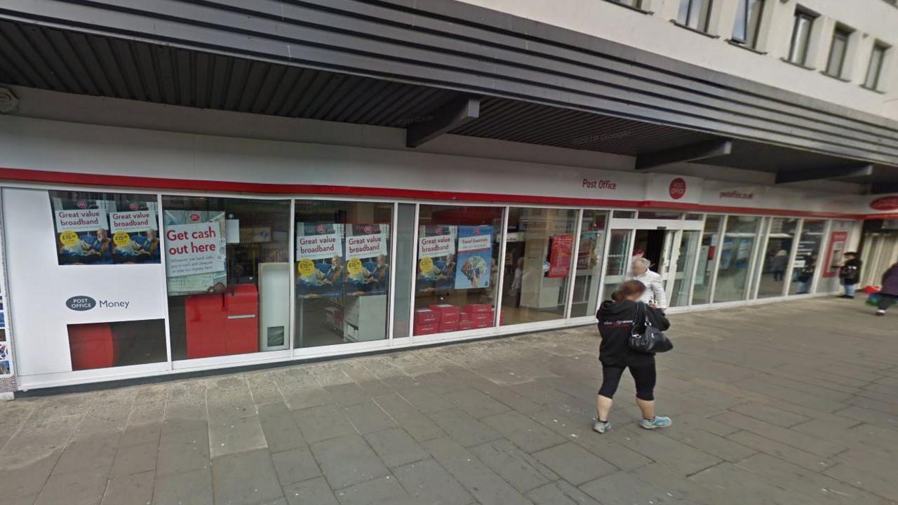 The Fawcett Street branch of the Post Office. The building is white fronted with red Post Office signage. People are walking along the pavement outside the branch.