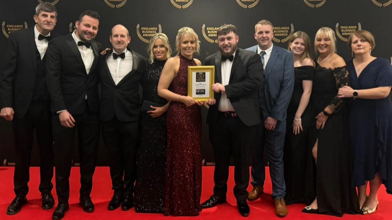 Ten people standing on a red carpet, posing for a photo. There are five men and five women. In the middle is Rachael who is blonde and wearing a red sparkly dress. She is holding a gold frame with an award inside it. Also holding the award is Joe, who is wearing a black and white suit and bow tie, and has brown hair and facial hair.