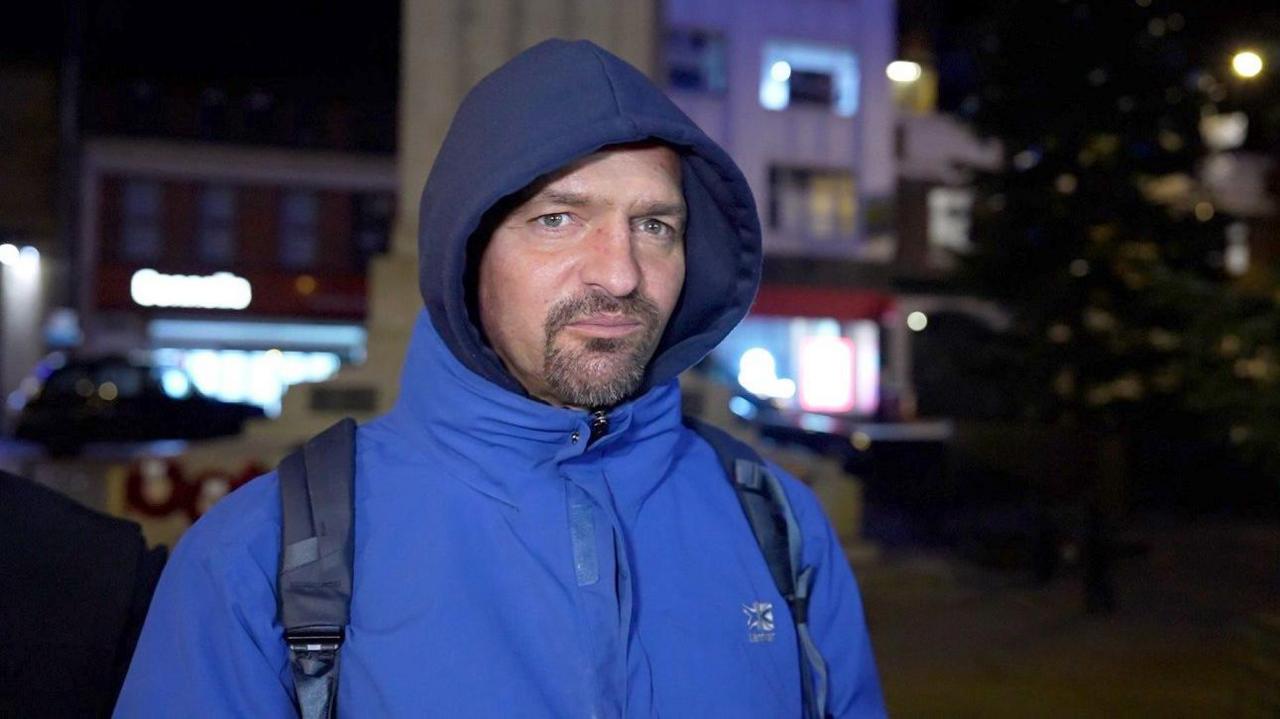 Nikolasz from Hungary looks at the camera, standing in Luton town centre with shops behind him. It is dark and there is the shape of a Christmas tree in the background. He wears a bright blue anorak over a dark blue hoodie, with his hood up. He has the straps of a blue rucksack over his shoulders. He is white with blue eyes and has a goatee beard.
