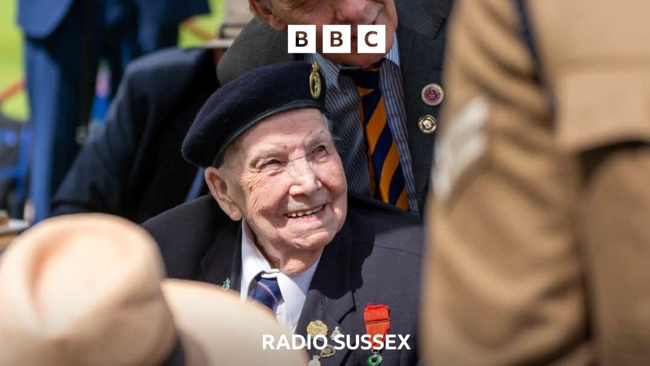 A World War 2 veteran smiles in the sunshine. He wears a blue uniform beret and his medals are pinned to his chest. He is surrounded by people.