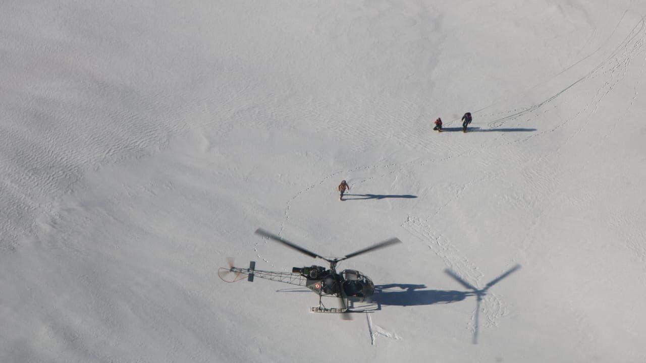 An aerial shot of a helicopter having landed on snow near two people with a third person walking between them