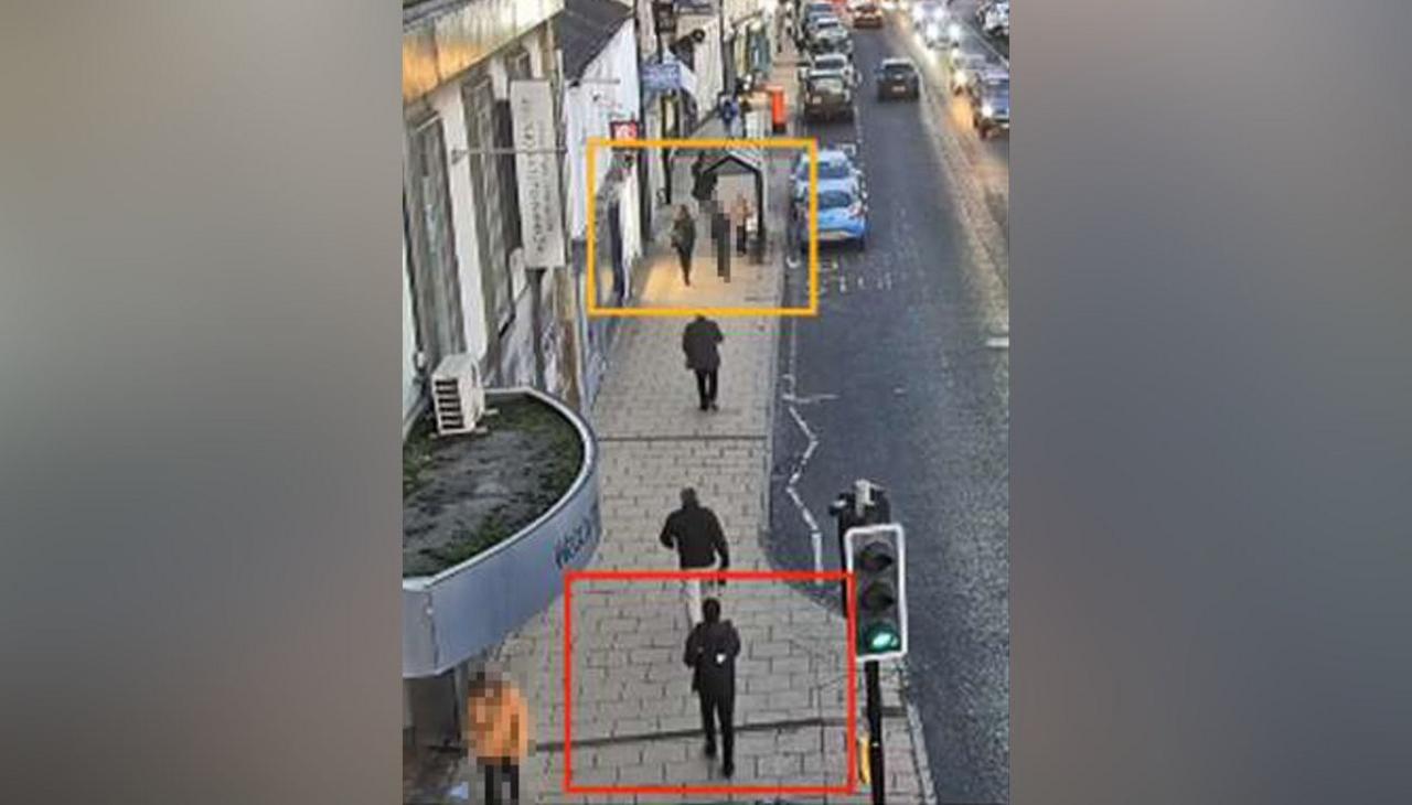 Still from CCTV showing people walking along a pavement on a shopping street. A figure all in black has been framed with a red box at the bottom of the street and a girl further ahead of him has had a yellow frame drawn around her