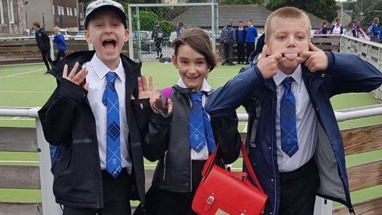 Aaron and Ryan in their school uniforms in the school playground pull silly faces for the camera. A female friend smiles in between the two of them. Their school ties are a light blue with a dark blue check pattern. The girl carries a bright red satchel.