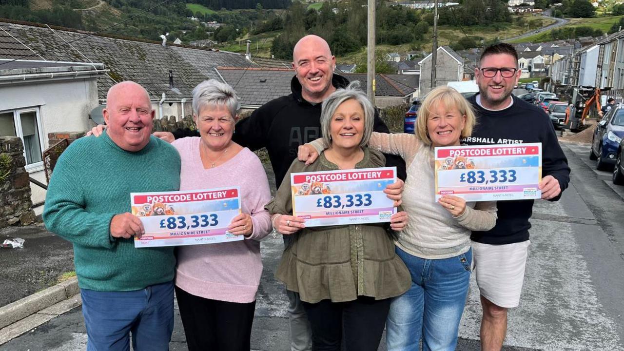 Six people stood on the street holding their winning cheques from the Postcode Lottery. The amount they have won, which is £83,333, is on the checks.