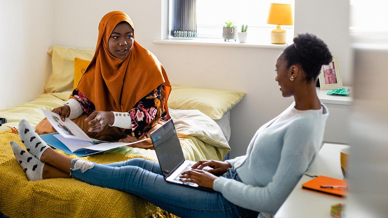 Two university students chat in a student dorm room, one is flicking through notes inside a blue ring binder, and the other is typing on a laptop on her lap.