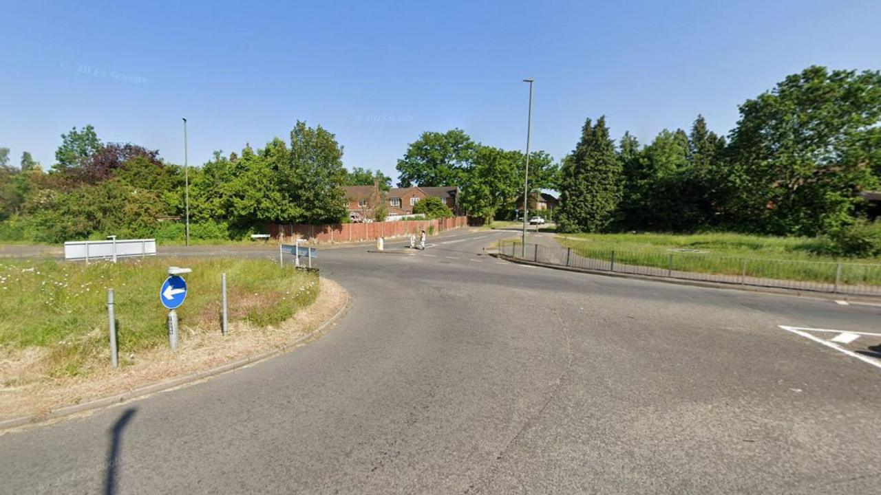 A Google Maps image of a roundabout. There is an area of grass on either side of the road. 