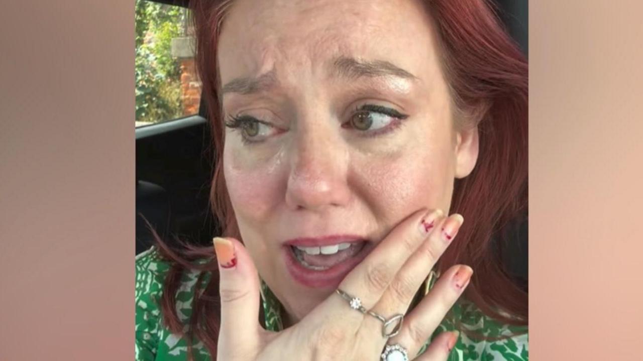 A red-haired woman with tears around her eyes and her hand up to her face