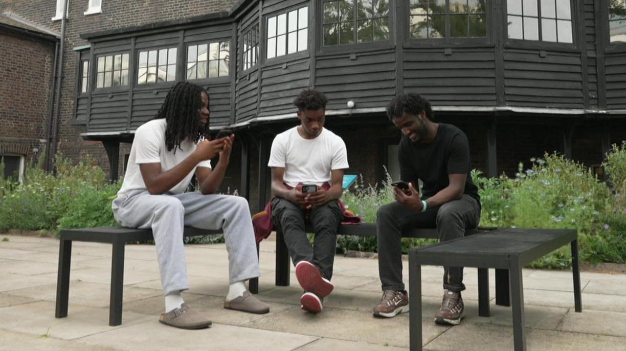 Prince, Henry and Adnan are sitting on some benches, in the gardens of the Museum of the Home. They are casually dressed and smiling as they chat and share some content on their mobile phones. 