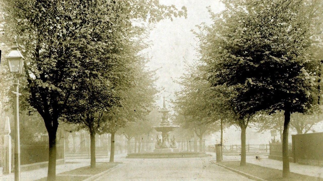 A sepia photograph of a tree-lined road with an ornate fountain in the middle