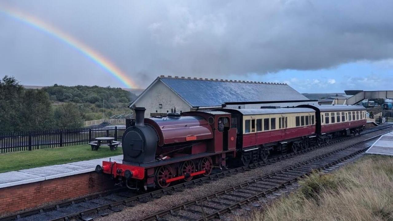 A red and cream train with an engine cabin and two passenger cabins. It's pulled in at a railway station near Blaenavon. There's a rainbow passing through the sky in the background, which is a mix of grey clouds and blue skies.