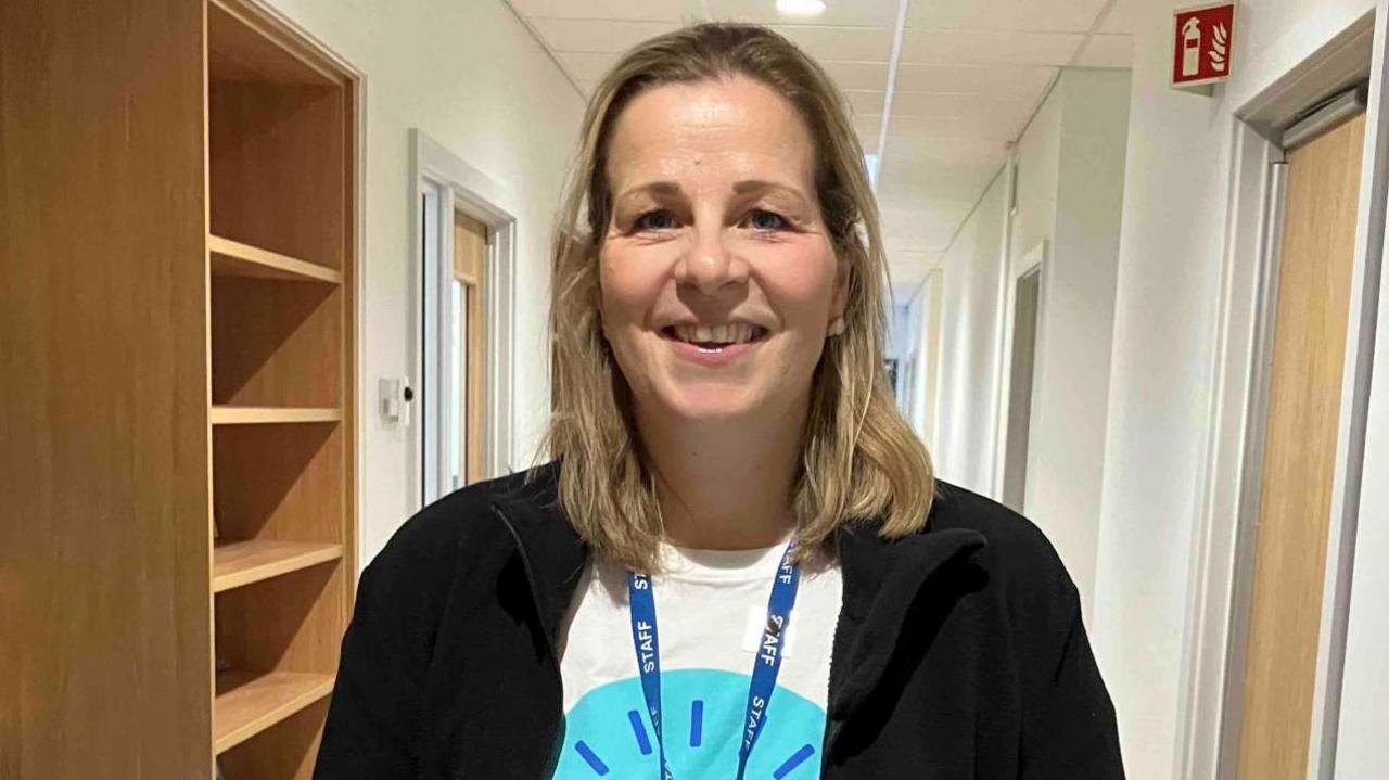 Image of a woman who is smiling at the camera. She has shoulder-length blonde hair, and is wearing a T-shirt with a blue design on the front, a blue staff lanyard and a black cardigan. In the background can be seen an empty bookshelf, a fire extinguisher sign and a number of internal doors.