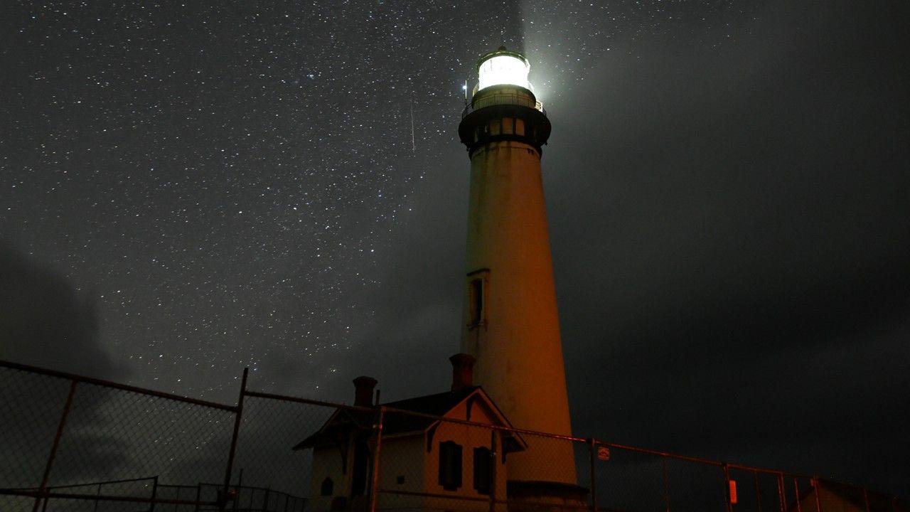 a light station with a small white house next to it, a railing behind it, under a dark grey and black night sky dotted with white starts and streaks of a meteor shower