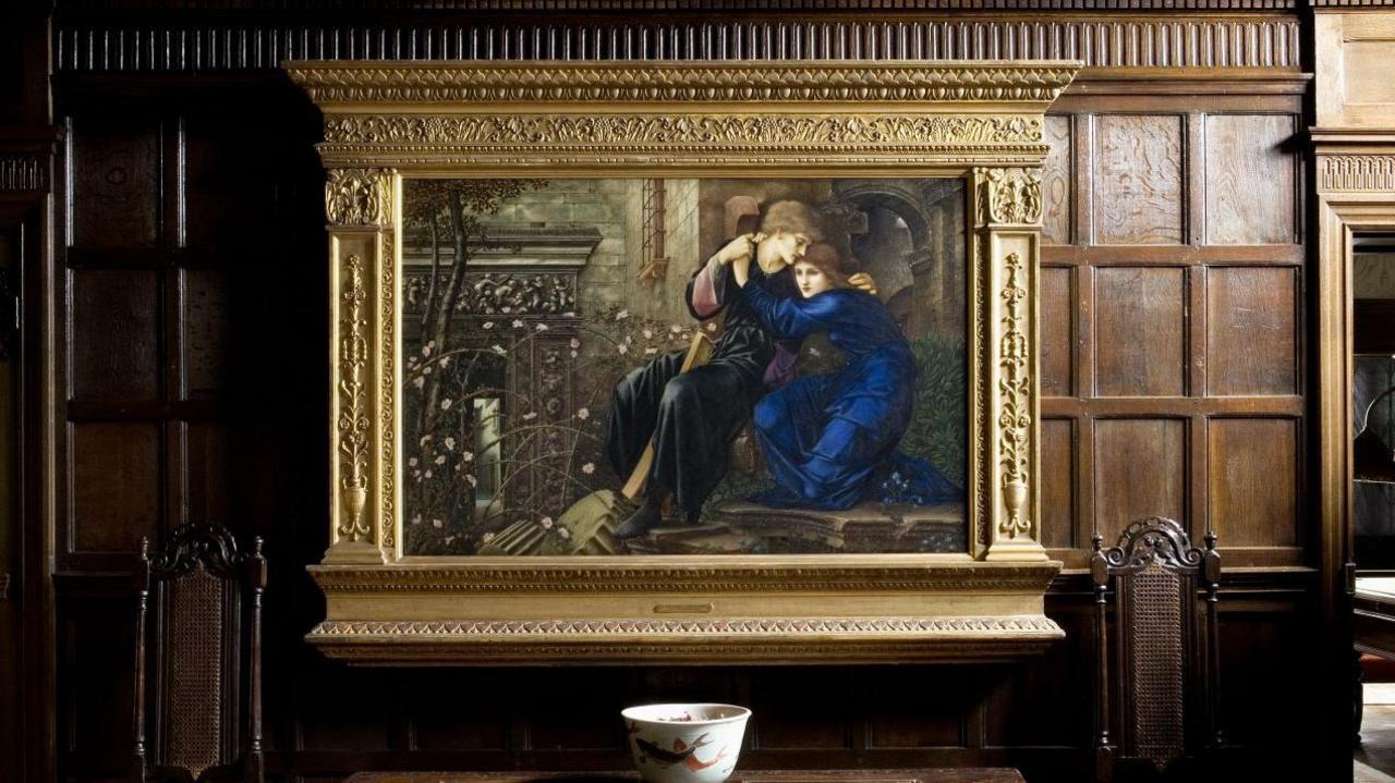 An oil painting in a gold frame hangs on a wall. The wall is made of dark wood. The painting shows a man and a woman wearing long robes and with their arms around each other.