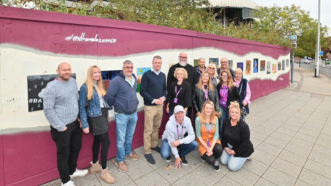 Fifteen people stood in front of a wall, facing the camera and smiling. Three of them are kneeling on the grey concrete and the rest are standing up. The wall is painted pink with a cream line in the middle. "#endhomelessness" is written in white writing at the top right of the art installation. There are paintings going along the cream part of the wall.