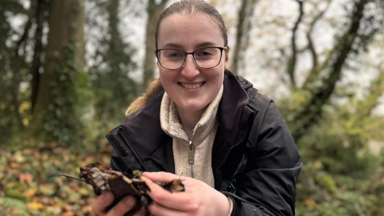 Niamh Carmichael, a woman with fair hair, glasses, dark jacket and brown fleece, holding leaves in her hands.
