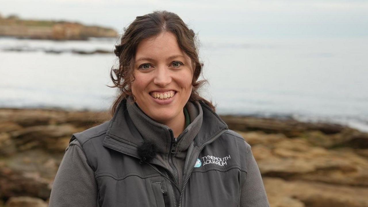 Ashleigh Bell, who is standing by the sea. She has brown curly hair, tied back in a pony tail, and is smiling broadly. She is wearing a grey fleece and bodywarmer with the words "Tynemouth Aquarium" on it.