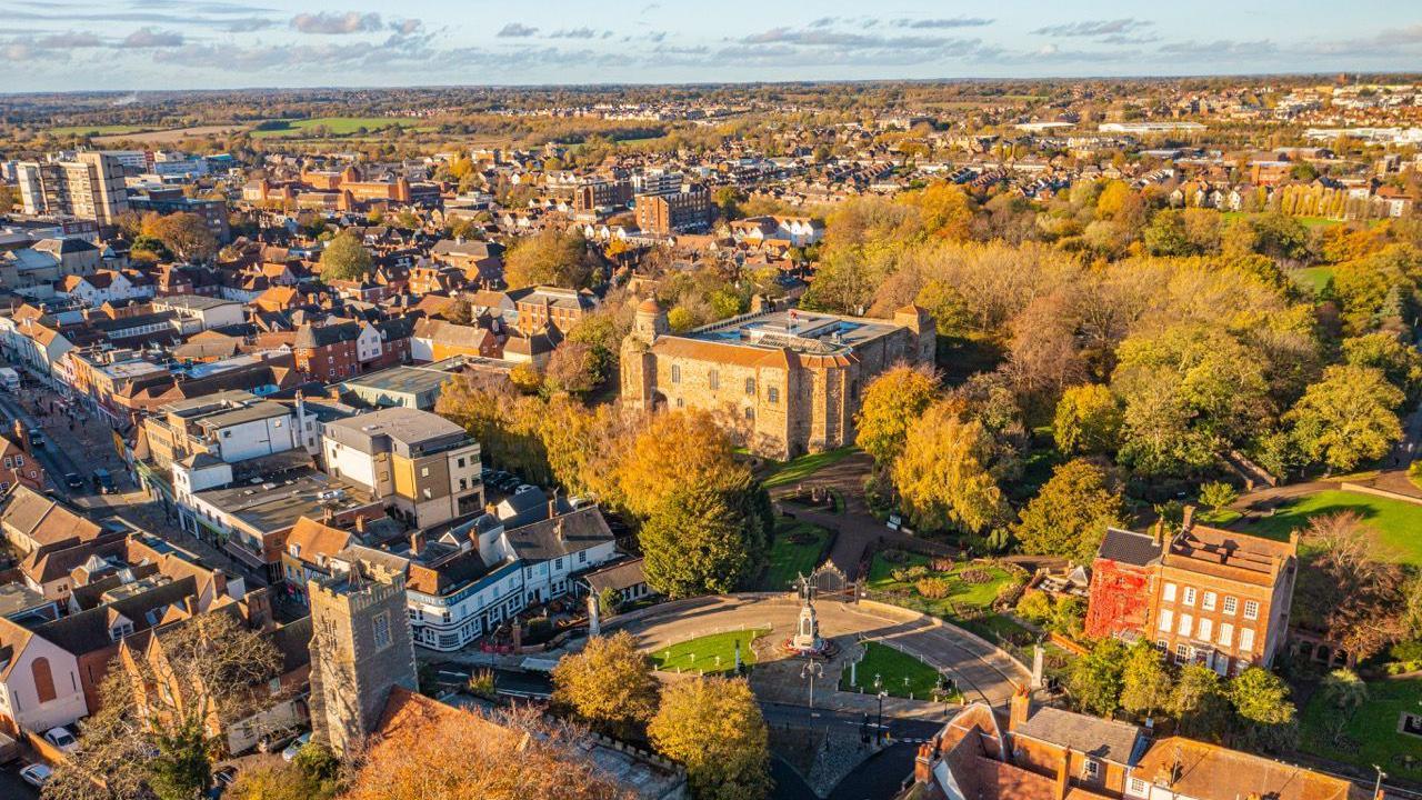 An aerial shot of Colchester with the castle in the centre of the picture.  There is trees of a park surroudning it and housing and some shops in the distance. The sun light is slowly setting on the city.