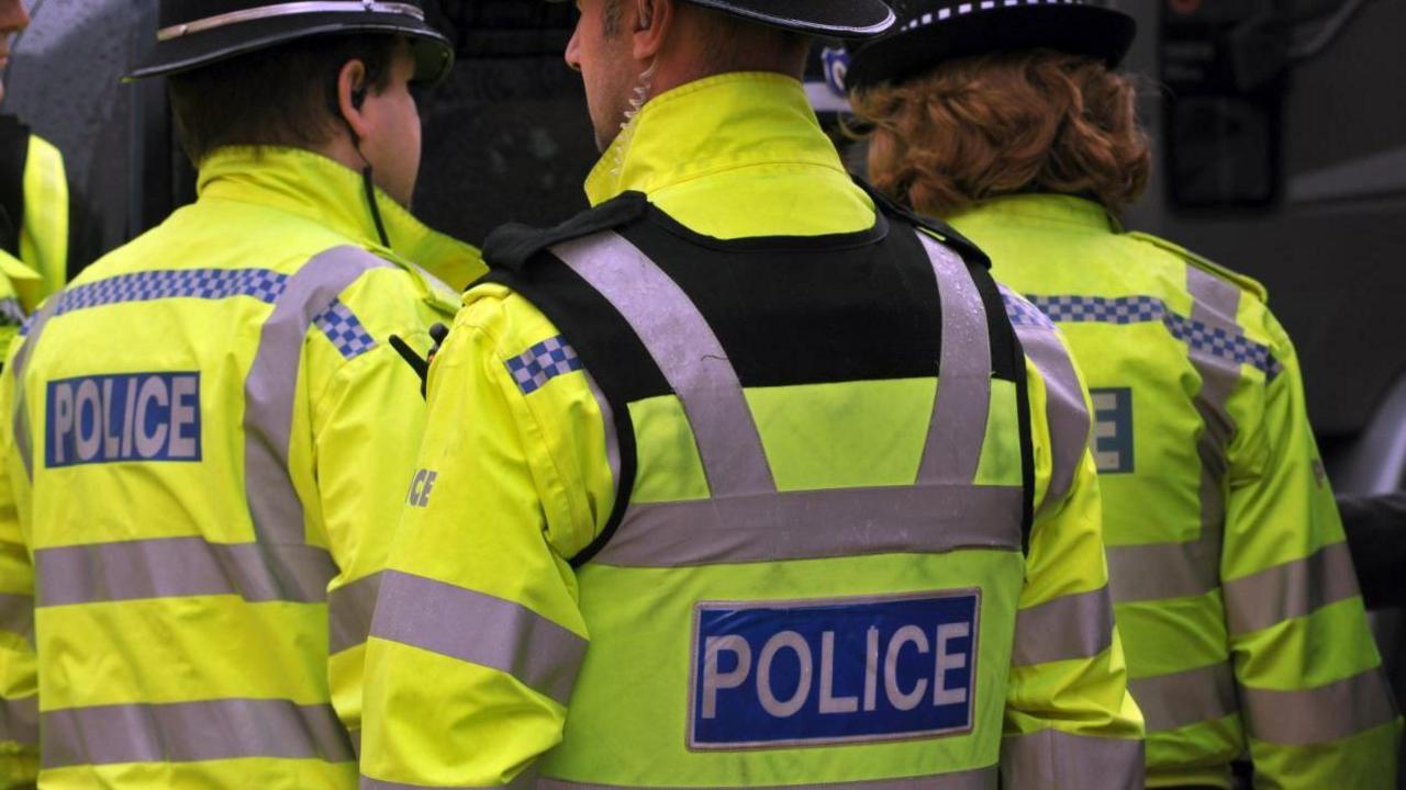 Three police officers wearing yellow high-vis jackets with 'police' written in white block capitals on a blue background at the scene of an incident.